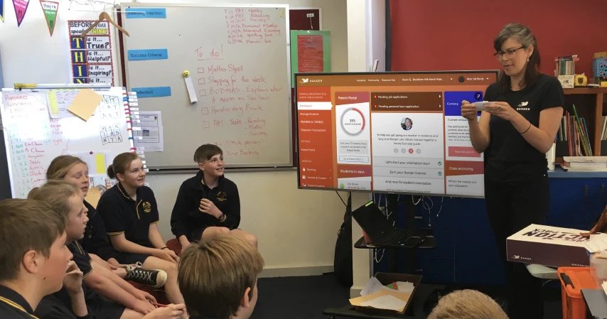 classroom with teacher presenting to students at the front of the class with banqer displaying on the screen behind her