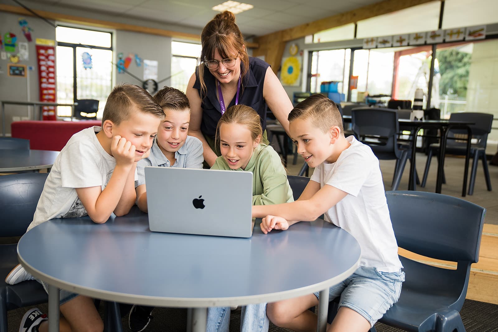 A group of four primary school students seated at a table and working together on a laptop. Behind them a teacher is supervising.