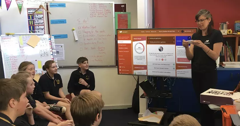 teacher addressing students in a classroom with banqer on the screen behind her