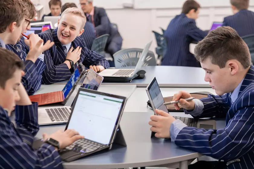 male students in a classroom on their laptops. one student is looking at a classmate and smiling